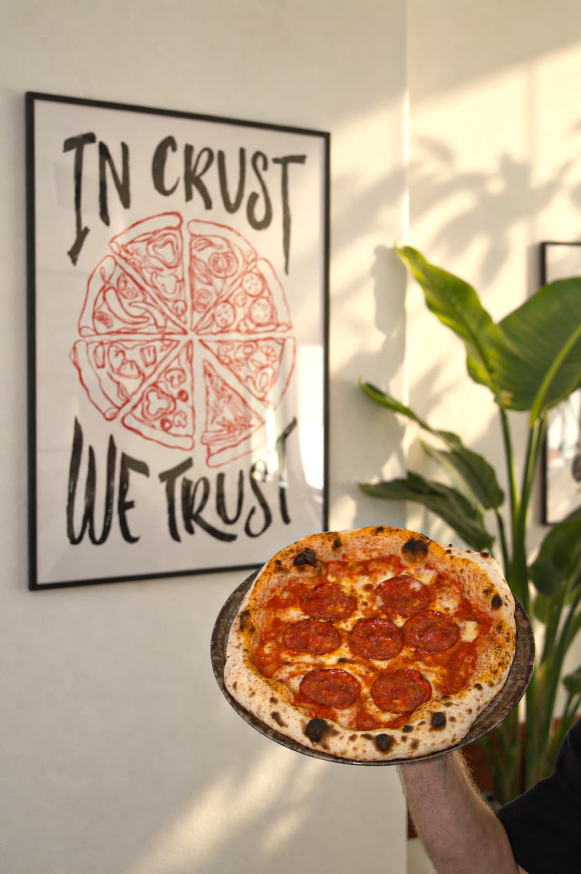 A hand holding a finished pizza in front of an In Crust We Trust poster